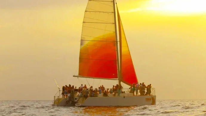 The catamaran under a red and orange sail against a golden sunset